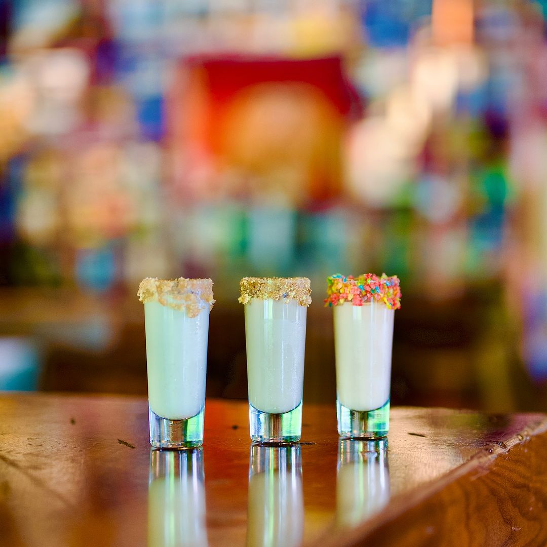 Three colorful shots lined up on a bar with cereal on their rims.